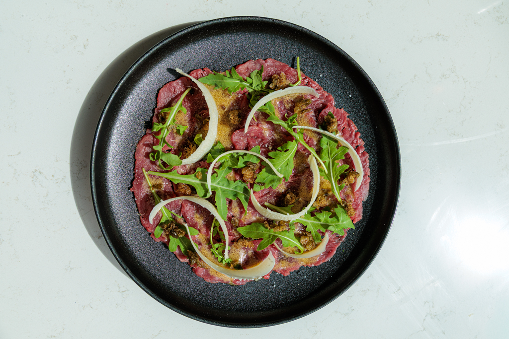 Carpaccio recept van diamanthaas, hand gesneden Dit carpaccio recept draait om techniek en kwaliteit. Diamanthaas een uur in de vriezer zodat het vlees stevig genoeg is om flinterdun te snijden. Dan met een scherp mes, plak voor plak, direct op koude borden. Vinaigrette van olijfolie, citroensap, Dijon en rode wijnazijn. Gefrituurde kappertjes die openbloemen in de olie. Parmezaan direct aan tafel geschaafd. Truffelolie op het allerlaatste moment. Wat maakt dit carpaccio recept anders? Hand gesneden geeft een heel andere textuur dan voorgesneden carpaccio. De plakken zijn dikker en malser, met meer bite en meer smaak. De kappertjes frituur je in neutrale olie tot ze openbloemen en krokant worden, dat geeft een textuur en intensiteit die je van rauwe kappertjes nooit haalt. En de truffelolie breng je pas op het allerlaatste moment aan, niet eerder, zodat het aroma niet verdampt voor het bord op tafel staat. Ingrediënten voor 4 personen 400 g diamanthaas, in één stuk 4 el olijfolie extra vergine sap van een halve citroen 1 tl Dijon mosterd 1 tl rode wijnazijn 50 g parmezaan, dungeschaafd 3 el kappertjes, goed drooggedept handvol rucola fleur de sel zwarte peper truffelolie voor de finishing neutrale olie om te frituren Bereiding Stap 1: Vlees invriezen Wikkel de diamanthaas strak in plasticfolie en leg 1 uur in de vriezer tot hij stevig maar niet bevroren is. Stap 2: Kappertjes frituren Dep de kappertjes volledig droog met keukenpapier. Verhit een laagje neutrale olie in een kleine pan op hoog vuur. Frituur de kappertjes 1 tot 2 minuten tot ze openbloemen en krokant zijn. Haal eruit met een schuimspaan en laat uitlekken op keukenpapier. Stap 3: Vinaigrette Meng olijfolie, citroensap, mosterd en rode wijnazijn tot een gladde vinaigrette. Breng op smaak met zout en peper. Stap 4: Vlees snijden Haal het vlees uit de vriezer en snijd met een scherp mes in flinterdunne plakken. Leg direct op koude borden en dek de hele bodem. Stap 5: Opmaken en serveren Drizzle de vinaigrette royaal over het vlees. Verdeel de rucola erover en schaaf de parmezaan direct aan tafel over het bord. Strooi de gefrituurde kappertjes erover. Breng op smaak met fleur de sel en zwarte peper. Druppel op het allerlaatste moment een paar druppels truffelolie erover en serveer direct. Wijn-spijs Een frisse droge witte wijn past perfect bij de delicate smaak van de carpaccio. Denk aan een Soave Classico, een lichte Pinot Grigio van Alturis of een Vermentino. De mineraliteit en frisheid sluiten mooi aan bij de truffelolie en de parmezaan. Wil je meer klassieke Italiaanse technieken leren? Bekijk ons workshopaanbod of lees ons recept voor vitello tonnato. Recept ontwikkeld door Mark Bosman — Ottavi Smaakwinkel & Kookstudio, Amsterdam YOAST CHECKLIST Keyphrase in paginatitel: staat exact vooraan. Goed. Keyphrase in introductie: staat in de eerste zin. Goed. Keyphrase dichtheid: staat in intro en subkop. Voldoende. Keyphrase in meta description: staat exact in de meta description. Goed. Keyphrase in subkop: staat in "Wat maakt dit carpaccio recept anders?" Goed. Interne links: link naar workshoppagina en vitello tonnato staan in de tekst. Goed. Uitgaande link: voeg een externe link toe, bijvoorbeeld naar een artikel over de geschiedenis van carpaccio op een betrouwbare culinaire bron. Alt-tag afbeelding: carpaccio recept van diamanthaas hand gesneden